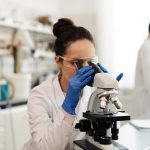 A female scientist in protective gear examines samples through a microscope in a laboratory setting. — BPC 157 10mg Vial for